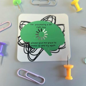 May include: A square white sticker with a green brain graphic and the text "I'm processing a lot right now, please give me grace to mess up and try again." The sticker is surrounded by push pins and paper clips.