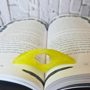 May include: A bright yellow, abstract-shaped bookmark rests on the open pages of a book. The bookmark has a circular cutout in the center and is made of a translucent material. The book's pages are white with black text.