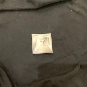 Puede incluir: Un objeto cuadrado de color plateado con un diseño de cuadrados anidados. El objeto está centrado en una superficie de tela oscura, posiblemente negra. El diseño consta de múltiples cuadrados concéntricos, creando un efecto de capas.