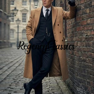 May include: A tan double-breasted overcoat worn over a black waistcoat, white collared shirt, black tie, and black trousers. The outfit includes a silver pocket watch chain attached to the waistcoat and black gloves. The text RegencyClassics is visible.