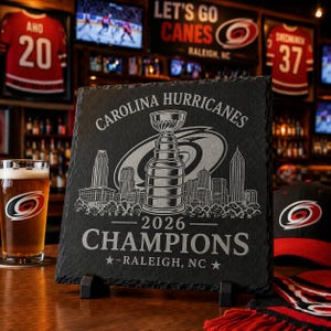 May include: A square black slate plaque featuring an engraved design of the Stanley Cup trophy centered over a city skyline. The text reads Carolina Hurricanes, 2026 Champions, and Raleigh, NC, with the team logo in the background.