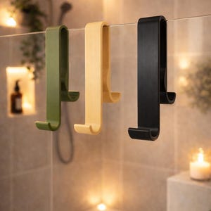 May include: Three shower door hooks in olive green, light yellow, and black hang on a glass shower door. The hooks have a curved design and are made of a matte material. The background is a tiled shower with soft lighting.