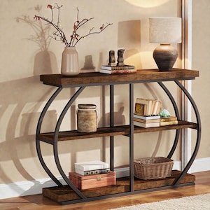 May include: A dark brown console table with three shelves and a curved, dark metal frame. The top shelf holds a lamp, vase, and books. The lower shelves display books, a basket, and a wooden box. The table is on a light-colored rug.
