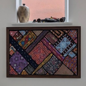 May include: A rectangular wall art piece featuring a patchwork of various colourful fabric swatches with intricate embroidery, small mirrors, and geometric patterns, all enclosed within a dark brown wooden frame and mounted on a plain white wall.