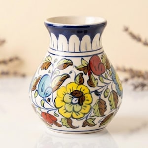 May include: A ceramic vase with a bulbous body and a flared rim, featuring a hand-painted floral pattern in yellow, blue, red, and green on a white background. The neck of the vase is decorated with a dark blue band and white petal motifs.