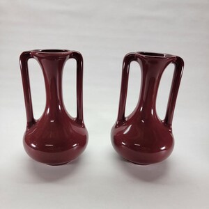 Può includere: Due vasi in ceramica color bordeaux scuro abbinati con finitura lucida. Ogni vaso presenta una base arrotondata, un collo sottile e due alti manici verticali che si estendono dal bordo fino alla spalla del recipiente, creando un aspetto simmetrico.