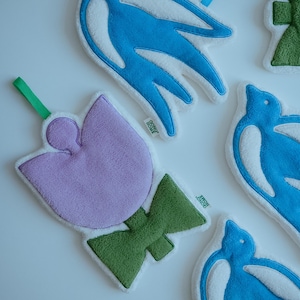 May include: A selection of soft, plush decorative items. The image shows a lavender tulip with a green stem and bow, and blue bird shapes with white detailing. Each item has a white border and a green loop.