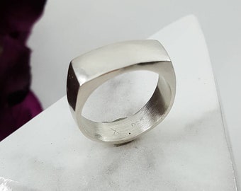 May include: A silver square band ring with a smooth, polished finish. The ring is displayed on a white marble-like surface.