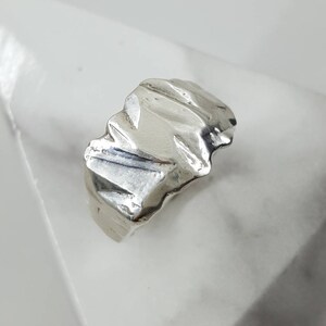 May include: A silver ring with a textured, abstract design. The ring is displayed on a white marble surface.