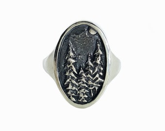 May include: A silver ring with an oval-shaped design featuring a detailed depiction of three pine trees under a crescent moon.