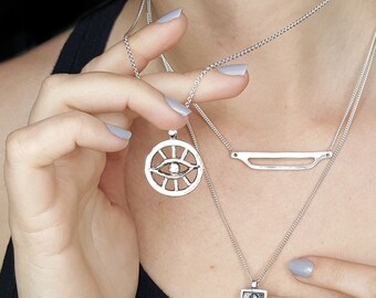 May include: Three silver necklaces with different pendants. The top necklace has a circular pendant with an eye design. The middle necklace has a thin, flat bar pendant. The bottom necklace has a rectangular pendant with a detailed image.