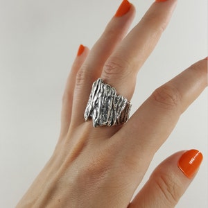 May include: A silver ring with a textured, organic design. The ring is wide and has a rough, natural look.