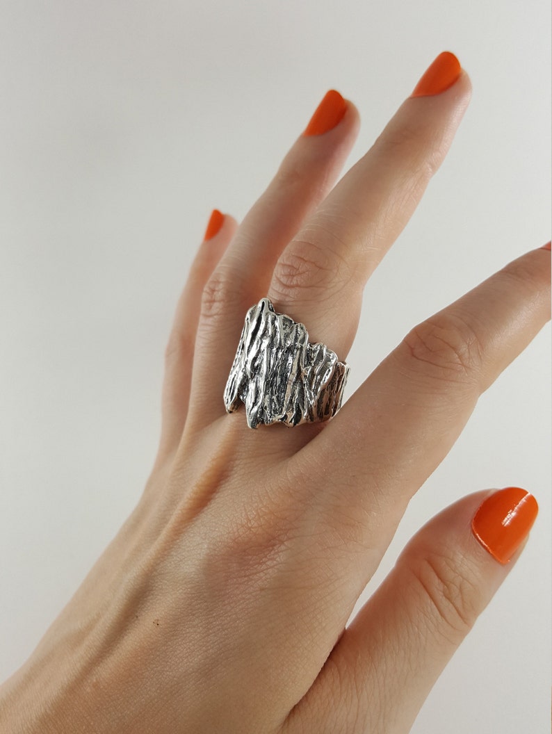 May include: A silver ring with a textured, organic design. The ring is wide and has a rough, natural look.