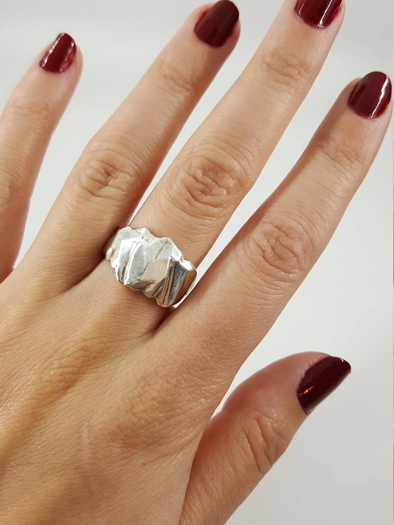 May include: A silver ring with a textured, wavy design. The ring is worn on a finger with dark red nail polish.