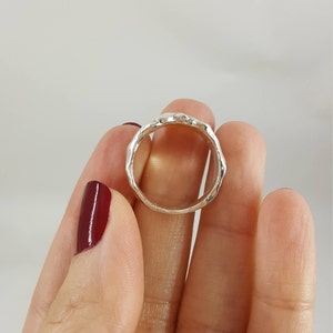 May include: A silver ring with a textured, hammered finish. The ring is shown on a finger.