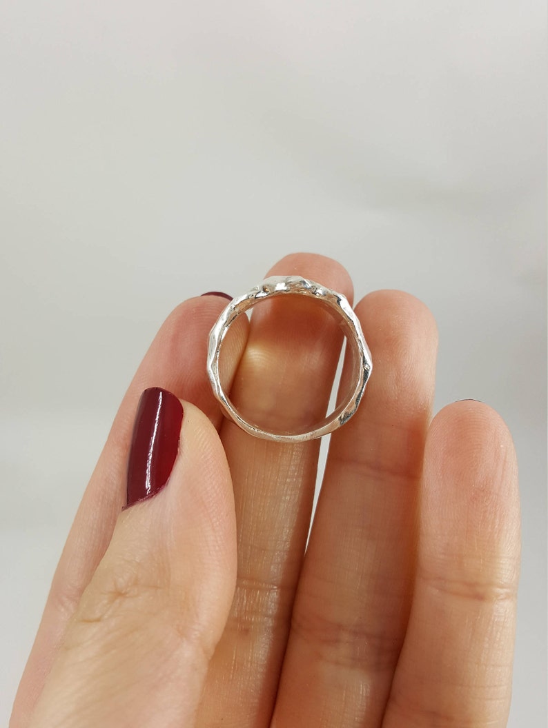 May include: A silver ring with a textured, hammered finish. The ring is shown on a finger.