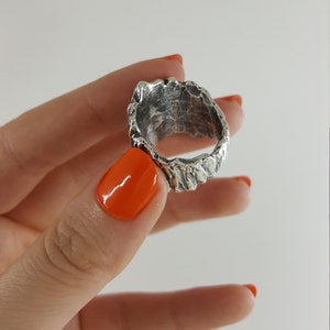 May include: A silver ring with a textured, organic design. The ring is shown on a finger with orange nail polish.
