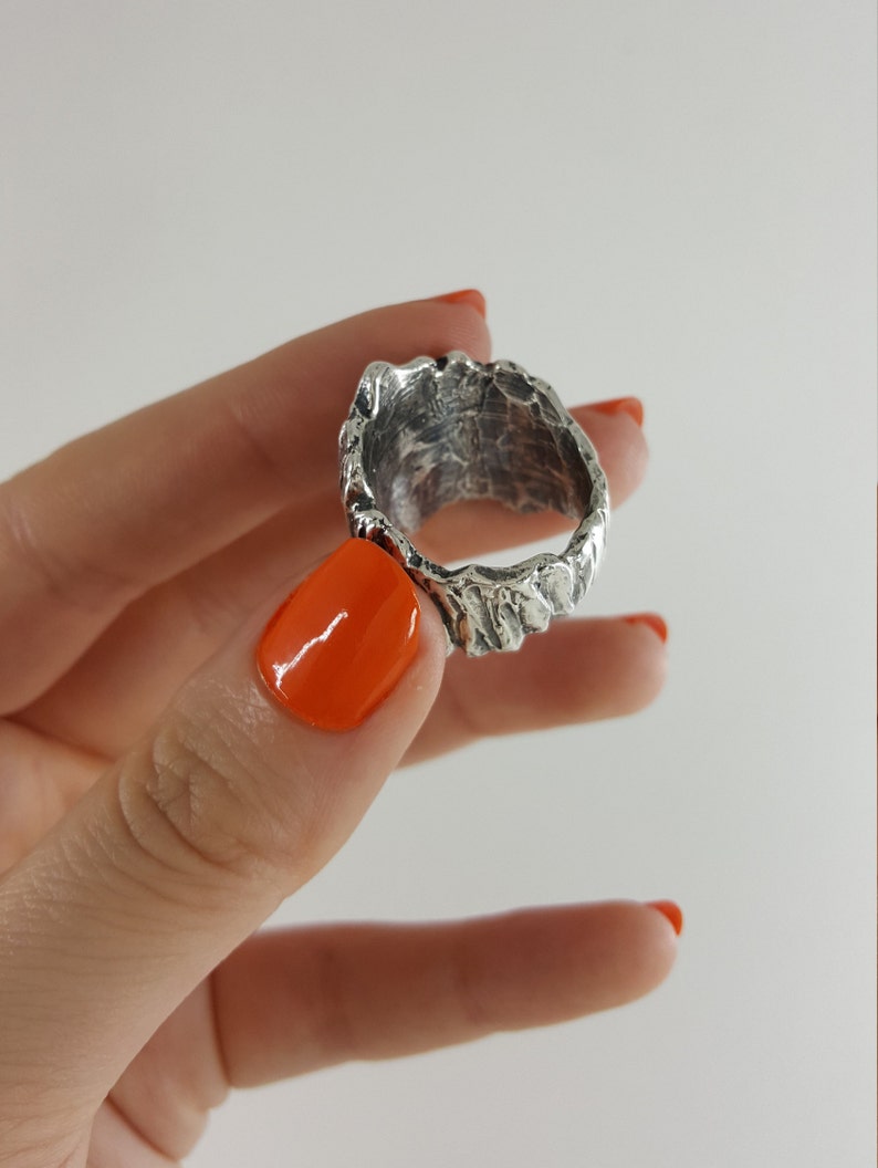 May include: A silver ring with a textured, organic design. The ring is shown on a finger with orange nail polish.