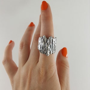 May include: A silver ring with a textured, organic design. The ring is worn on a finger with orange nail polish.