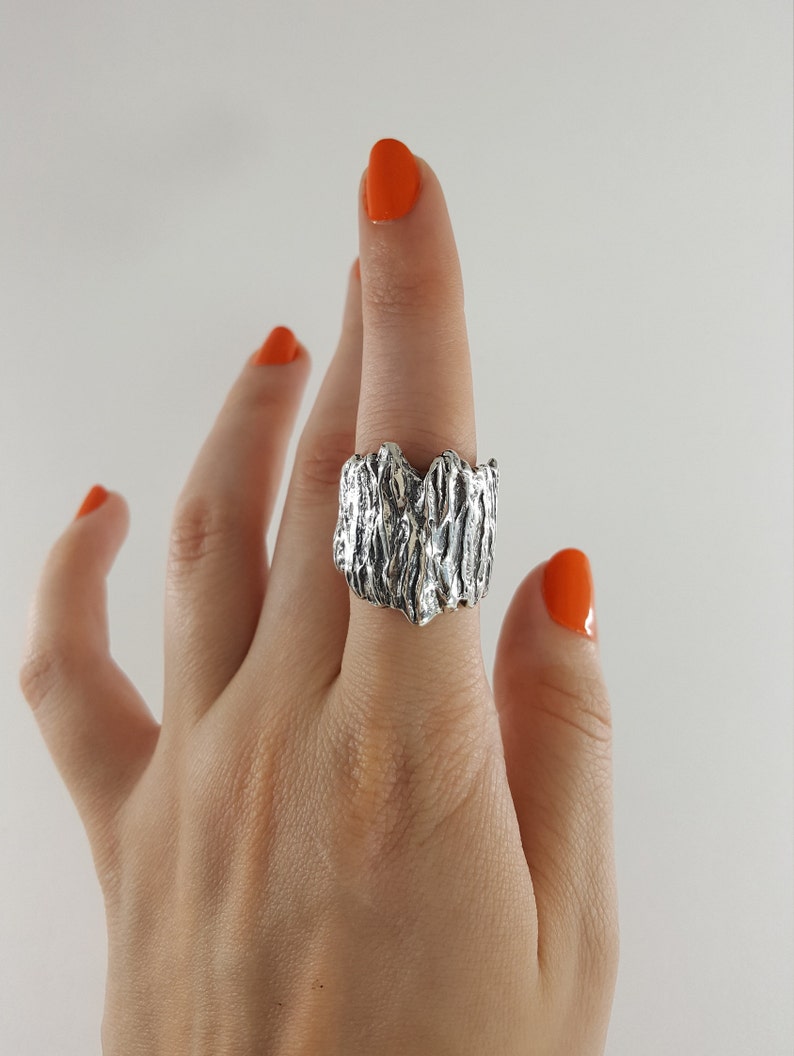 May include: A silver ring with a textured, organic design. The ring is worn on a finger with orange nail polish.
