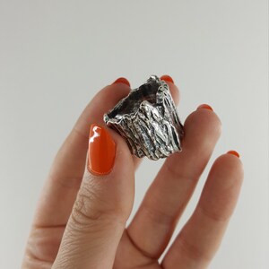 May include: A silver ring with a textured, tree bark-like design. The ring is shown on a hand with orange nail polish.