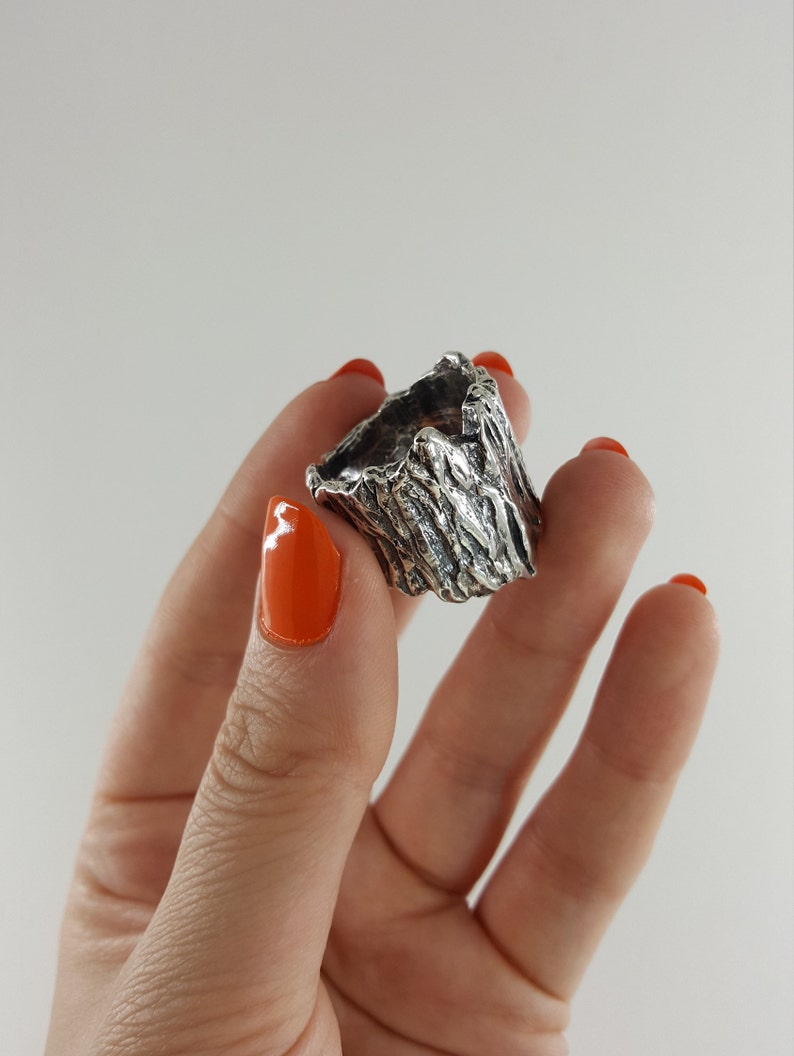 May include: A silver ring with a textured, tree bark-like design. The ring is shown on a hand with orange nail polish.