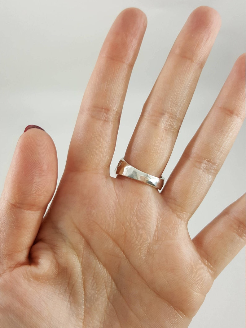 May include: A silver ring with a textured, hammered finish. The ring is worn on the middle finger of a hand.
