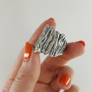 May include: A silver ring with a textured, organic design resembling a mountain range. The ring is worn on a finger with orange nail polish.