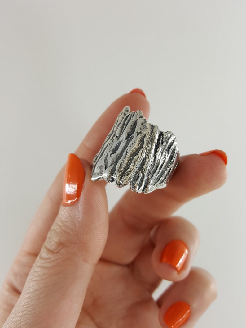 May include: A silver ring with a textured, organic design resembling a mountain range. The ring is worn on a finger with orange nail polish.