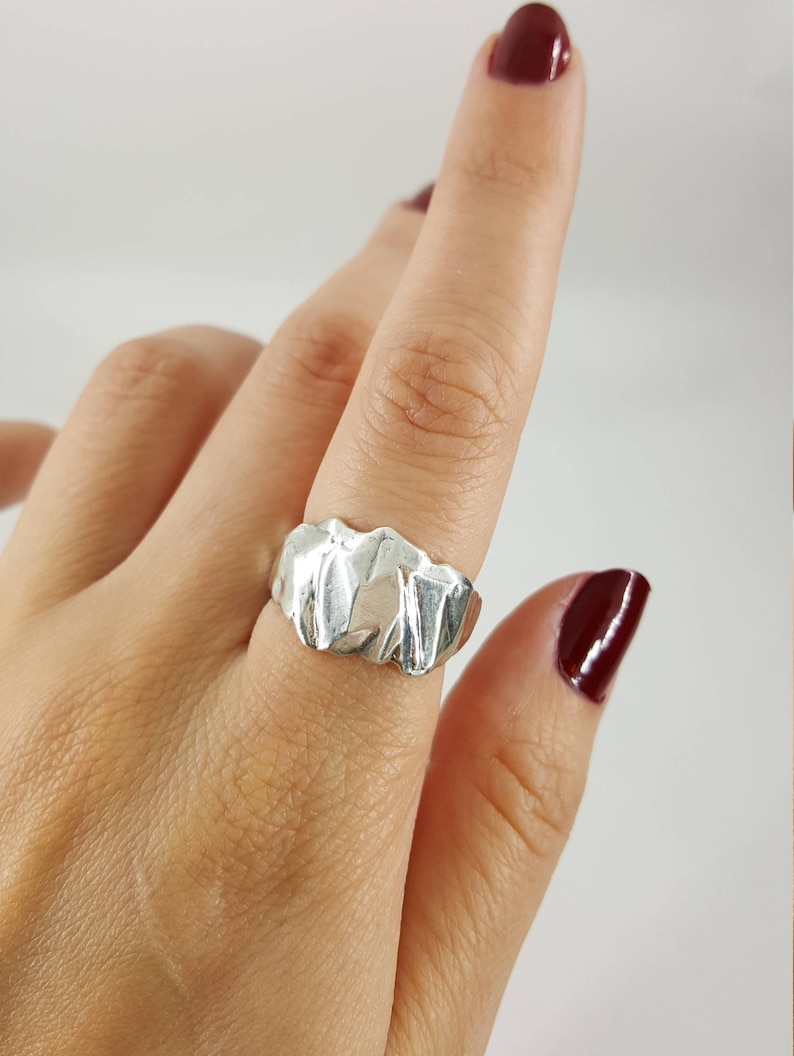 May include: A silver ring with a textured, abstract design. The ring is worn on a finger with red nail polish.
