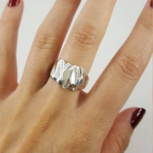 May include: A silver ring with a textured, abstract design. The ring is worn on a person's finger.