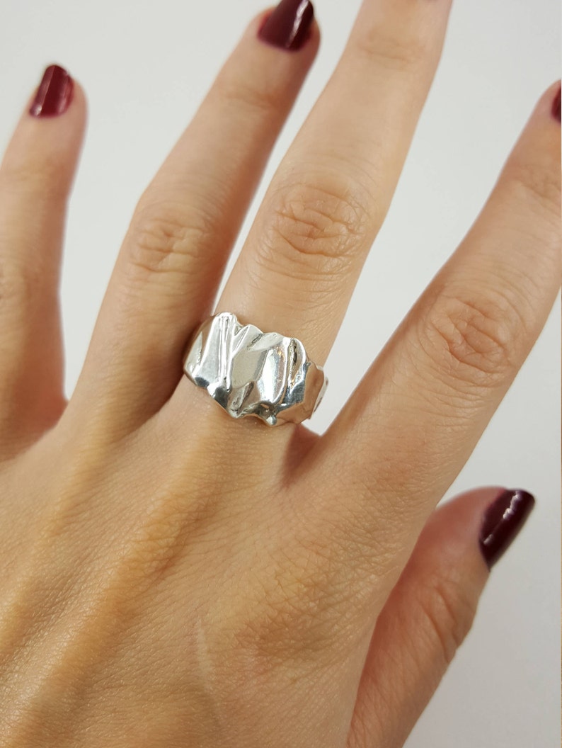 May include: A silver ring with a textured, abstract design. The ring is worn on a person's finger.