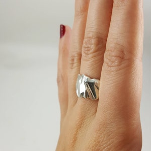 May include: A silver ring with a textured, organic design. The ring is worn on a finger with red nail polish.