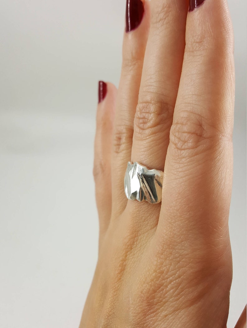 May include: A silver ring with a textured, organic design. The ring is worn on a finger with red nail polish.