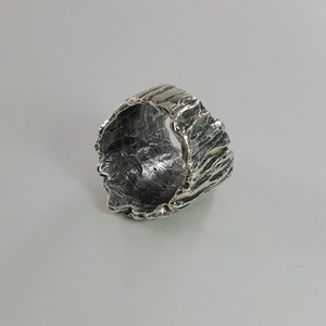 May include: A silver ring with a textured, organic design. The ring is wide and has a rough, uneven surface.