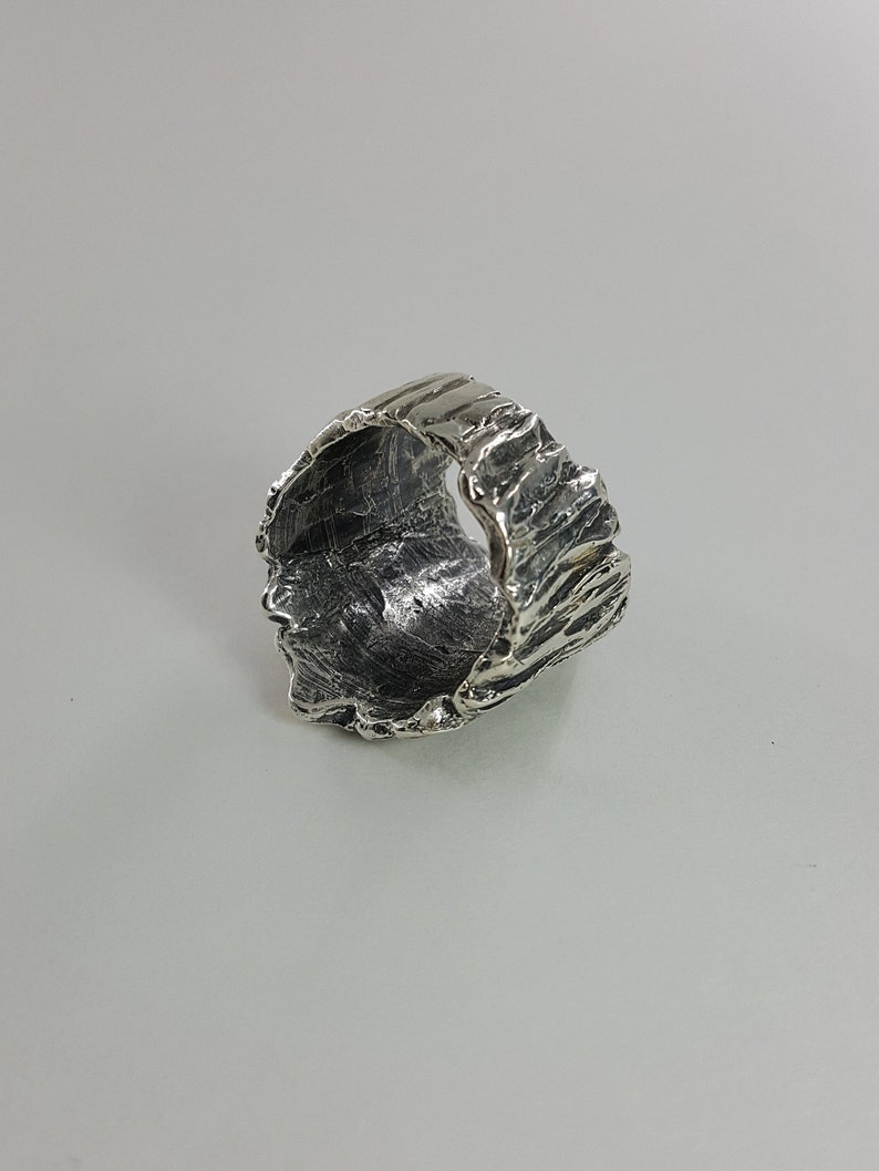 May include: A silver ring with a textured, organic design. The ring is wide and has a rough, uneven surface.
