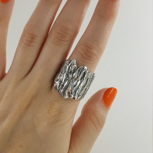 May include: A silver ring with a textured, organic design, resembling a piece of wood. The ring is worn on a hand with orange nail polish.