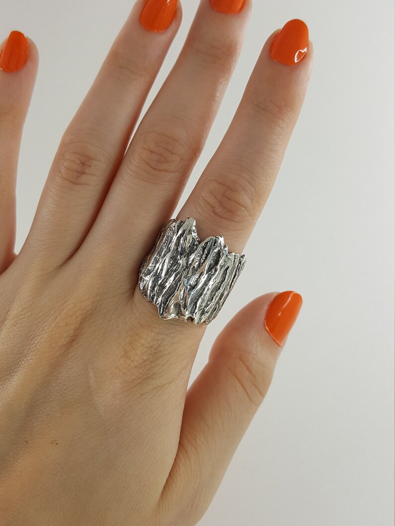 May include: A silver ring with a textured, organic design, resembling a piece of wood. The ring is worn on a hand with orange nail polish.