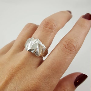 May include: A silver ring with a textured, abstract design. The ring is worn on a finger.