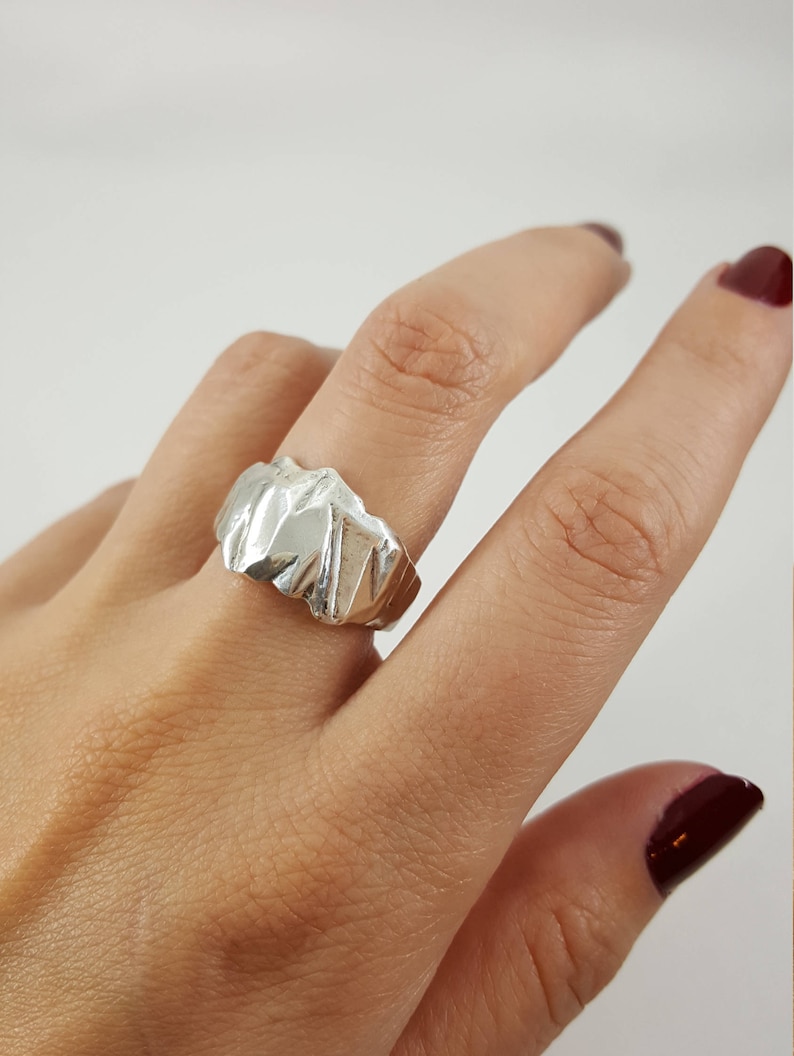 May include: A silver ring with a textured, abstract design. The ring is worn on a finger.
