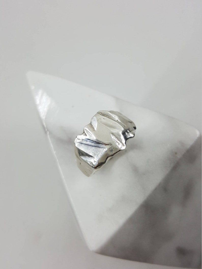 May include: A silver ring with a textured, abstract design. The ring is displayed on a white marble surface.