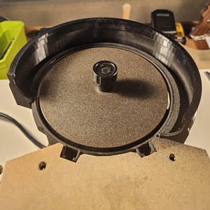 Puede incluir: Primer plano de una amoladora de banco con una muela de color marrón oscuro. La amoladora tiene una carcasa de plástico negro y una base de madera. La muela está rodeada por un protector negro. La imagen está bien iluminada.