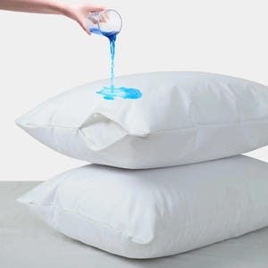May include: Two stacked white rectangular pillows with a zippered closure on the top pillow. A clear glass is pouring blue liquid onto the surface of the top pillow, where the liquid beads up on the fabric, demonstrating a water-resistant property.