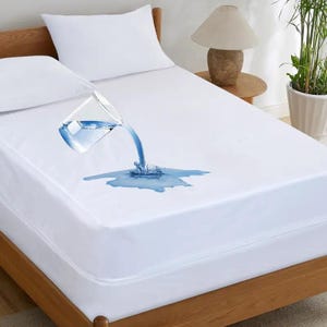 May include: A white waterproof mattress protector fitted over a bed with two white pillows. A clear glass pitcher is pouring water onto the surface, showing the liquid pooling on top of the fabric without soaking into the mattress protector.