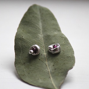 Puede incluir: Dos pendientes de plata con un diseño texturizado y orgánico, cada uno con una pequeña gema de color rojo oscuro. Los pendientes se muestran sobre una gran hoja verde, creando una presentación natural y artística. Joyas únicas.