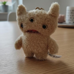 Peut inclure: Un jouet monstre en peluche beige avec trois yeux, deux petits bras et deux oreilles. Le jouet a une bouche rouge et une étiquette qui dit "MADE IN CHINA". Le jouet est posé sur une surface en bois.