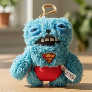 Peut inclure: Un jouet en peluche bleu avec le logo Superman sur la poitrine et un vêtement rouge en forme de cœur. Le jouet a un visage de monstre avec de grands yeux et un sourire édenté. Une étiquette indiquant "MADE IN CHINA" est attachée.