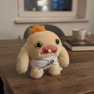 Peut inclure: Une peluche représentant une créature avec un corps beige clair, de grands yeux jaunes et une touffe de cheveux orange. Elle porte une couche blanche avec un motif noir. Le jouet est sur une table en bois brun.