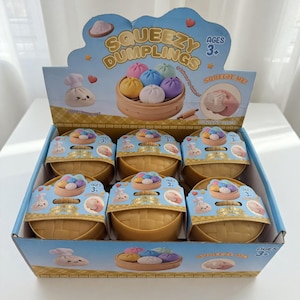 Puede incluir: Una caja expositora de juguetes Squeezy Dumplings. La caja presenta un fondo azul claro con las palabras "Squeezy Dumplings" y "Ages 3+". En el interior hay varios juguetes con forma de dumpling de varios colores, empaquetados en pequeños recipientes en forma de cesta.