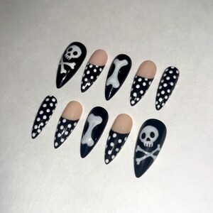 May include: A set of ten stiletto-shaped press-on nails featuring a black base with white polka dots, white skull and crossbones motifs, and white bone designs. Some nails have a nude-colored half-moon base, while others are fully patterned.